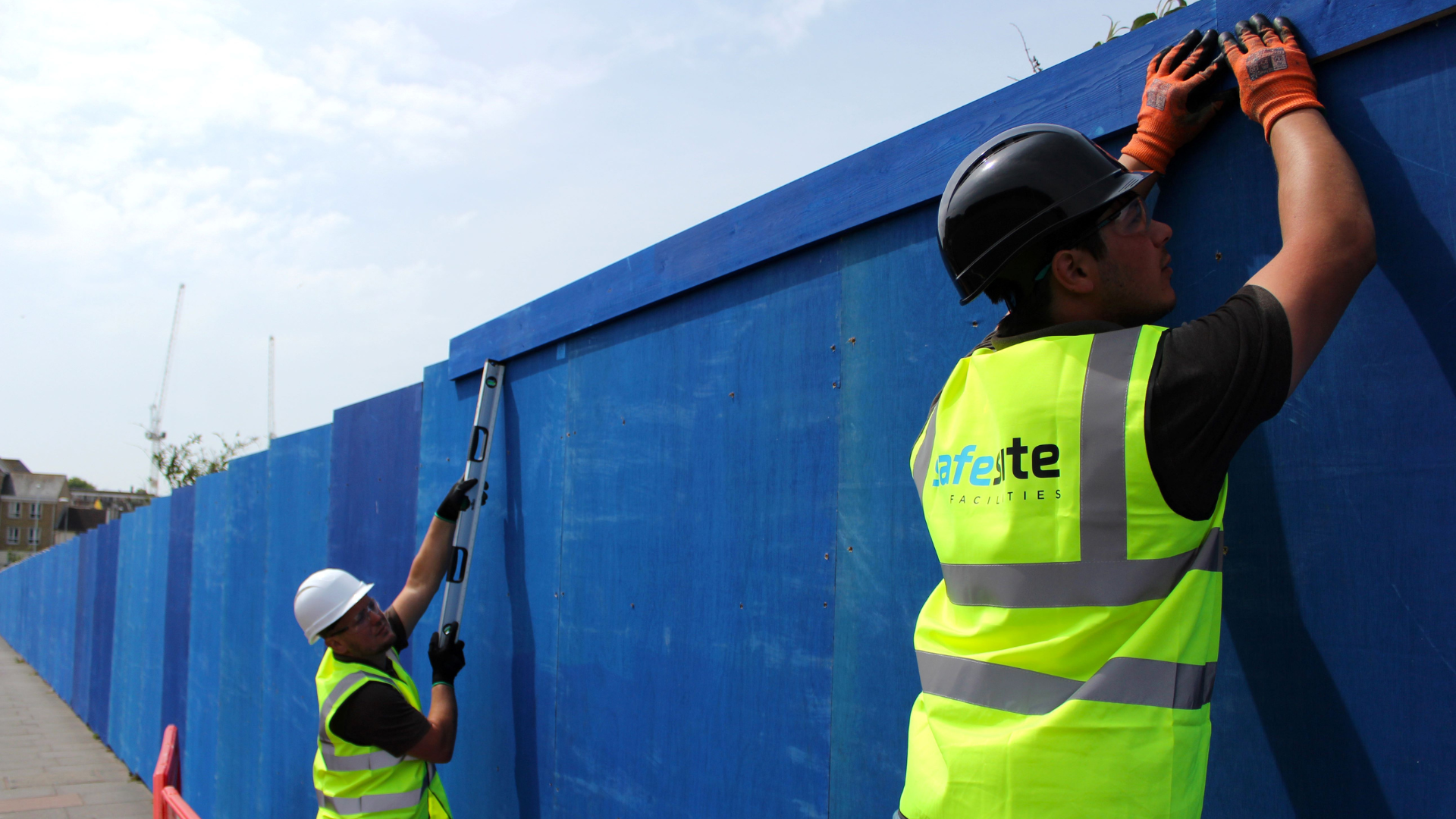 Two construction workers in safety gear are securing a blue fence. One holds a level while the other adjusts the top section.