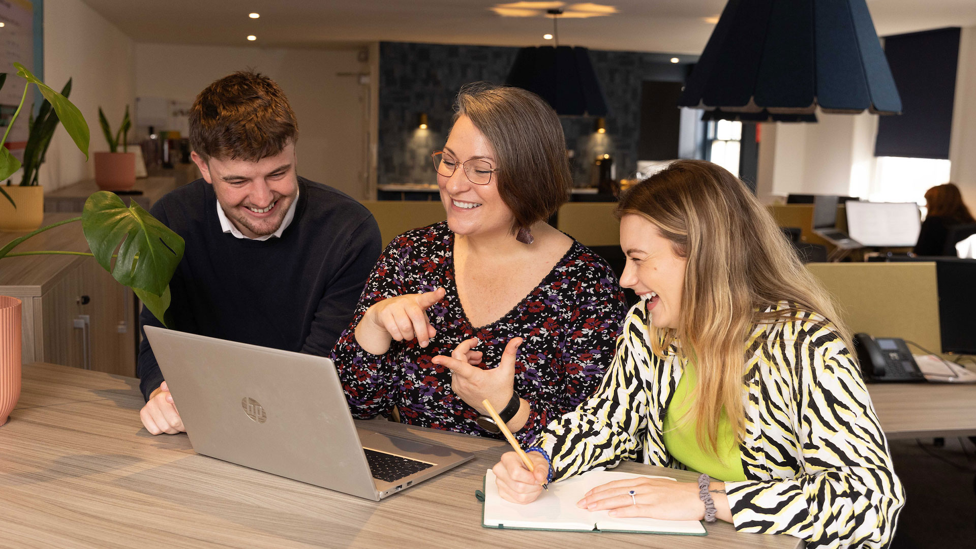 Three people are sitting at a table, looking at a laptop. One person is gesturing while others are smiling and taking notes in a brightly lit office space.