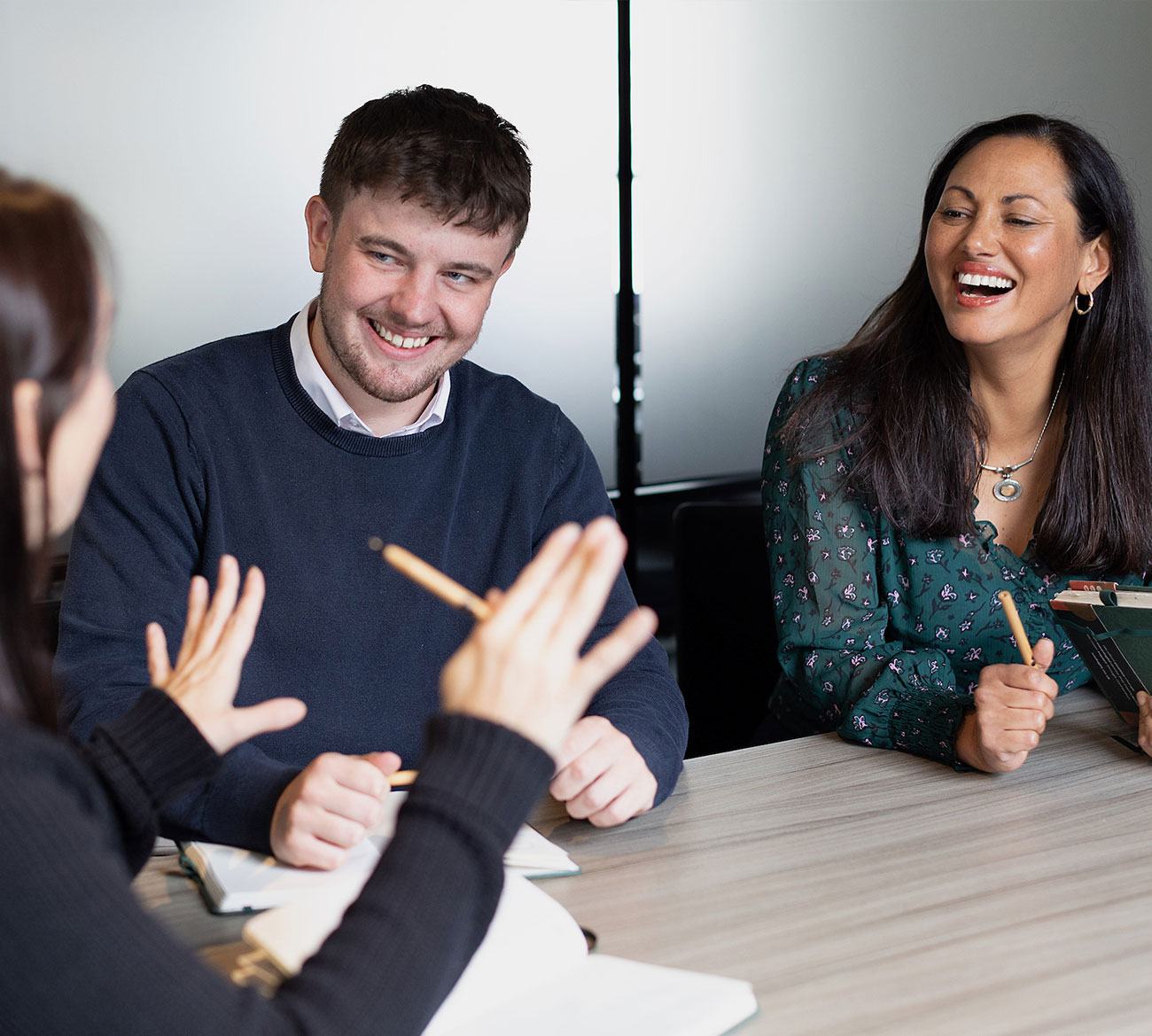 Three people sit at a table, engaged in a lively discussion with notebooks and pens, smiling and gesturing.