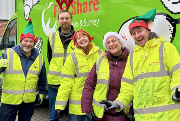 Five people wearing festive hats and high-visibility jackets smile and gesture in front of a green van with a "FareShare Sussex & Surrey" logo.